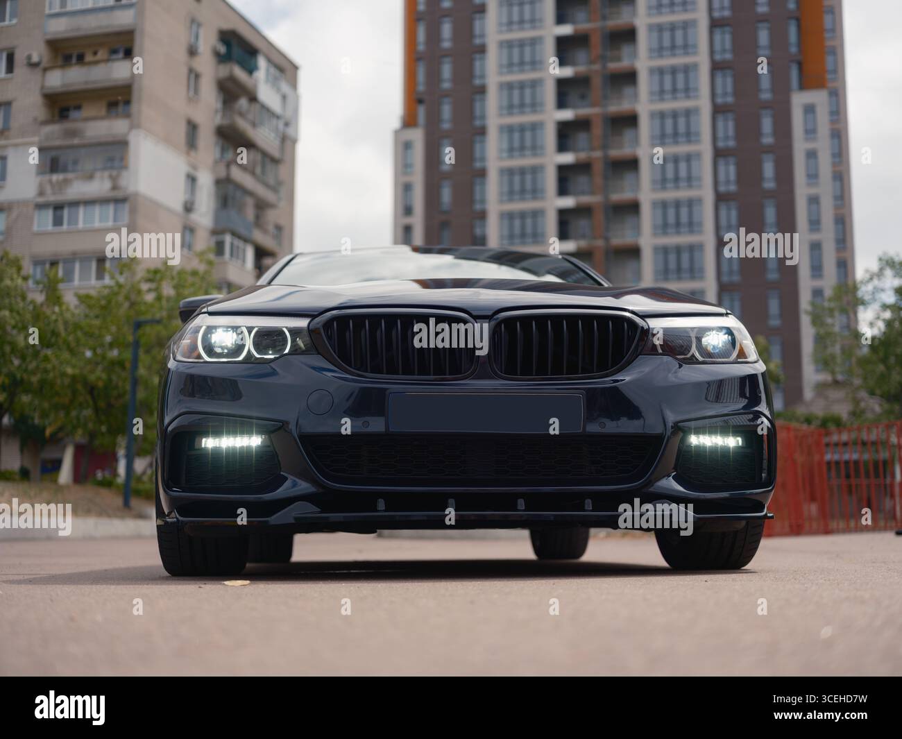 Front view dark sedan parked on city street. Modern design, sleek headlights, and urban buildings in background. Ideal for automotive and city life vi Stock Photo