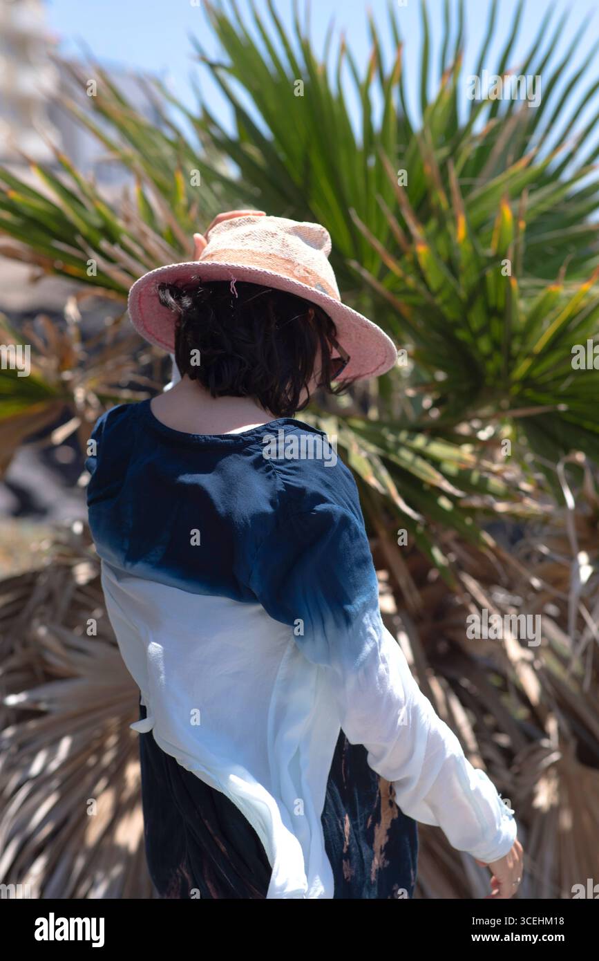Half-length portrait of a brunette woman wearing a hat and dark blue and white shirt, holding the hat with one hand. She stands in front of sunlit pal Stock Photo