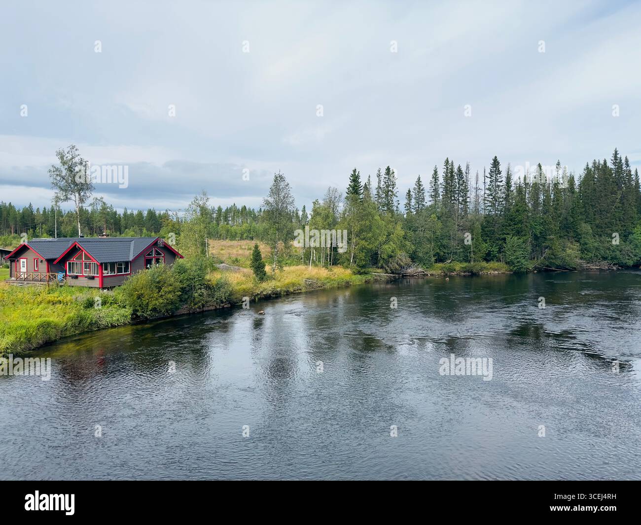 A picturesque red cabin by a flowing river, surrounded by lush greenery. Stock Photo