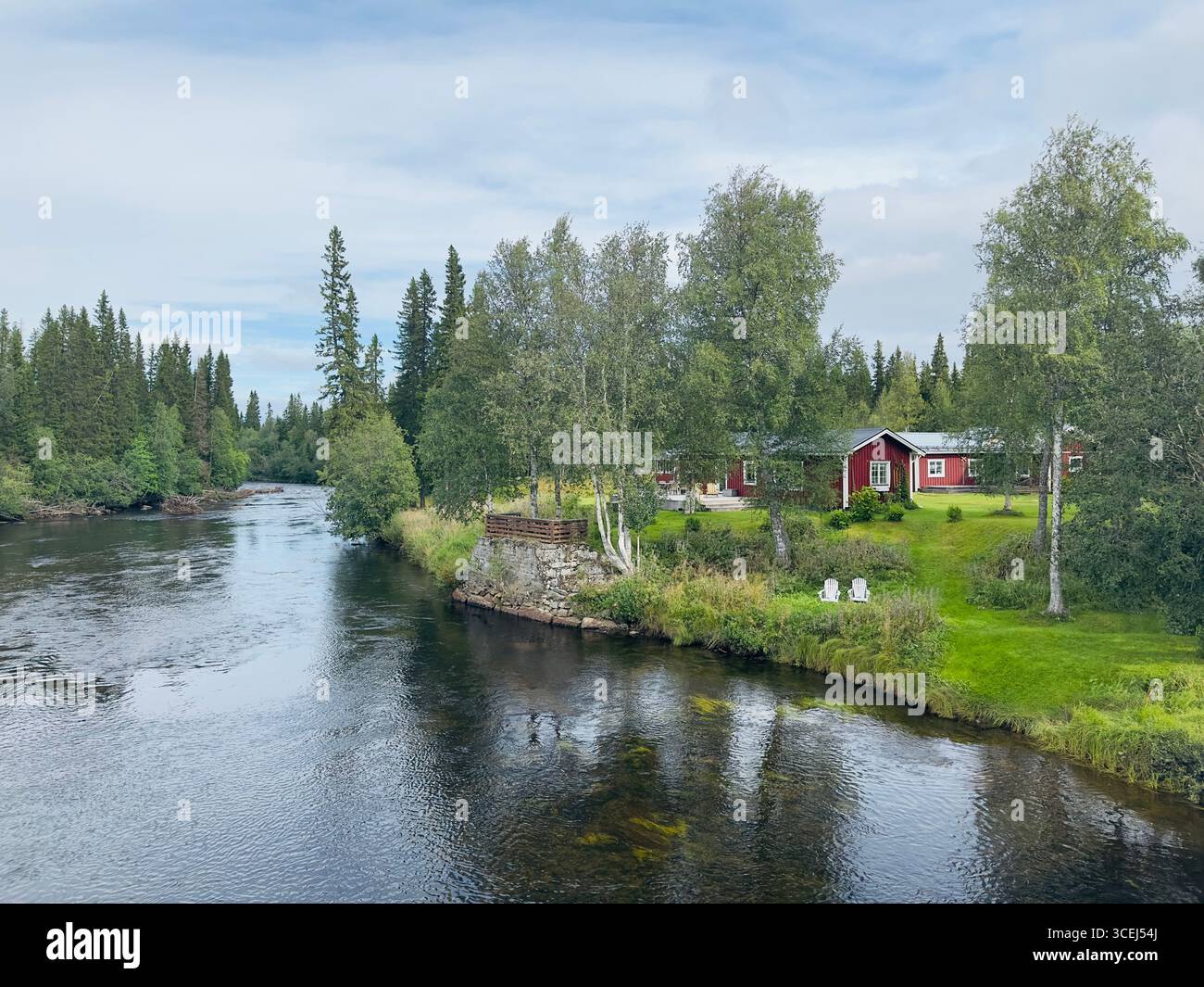 A picturesque red cabin by a flowing river, surrounded by lush greenery. Stock Photo