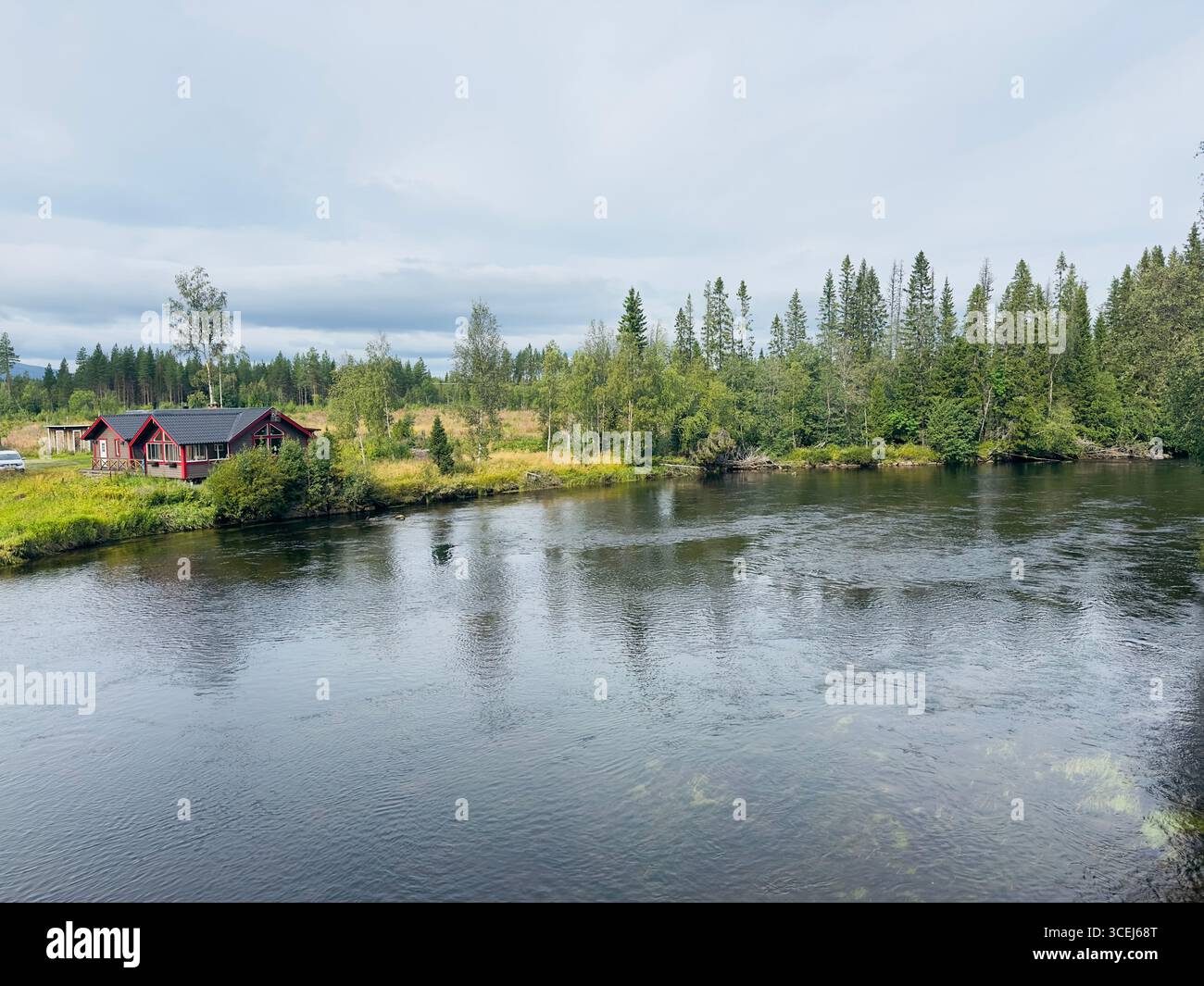 A red cabin sits peacefully on a riverbank, surrounded by lush green trees and calm water. Stock Photo