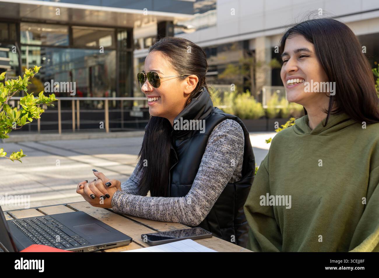 Two happy businesswomen are working remotely on a laptop while sitting at a table outside Stock Photo