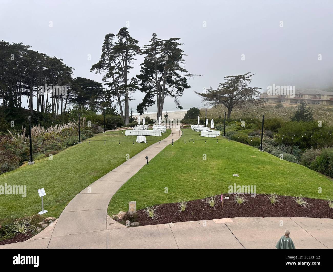 Elegant outdoor wedding and event setup with white tables, chairs, and umbrellas arranged on a lawn overlooking the Pacific Ocean on a foggy day - Smartphone Captured Stock Image