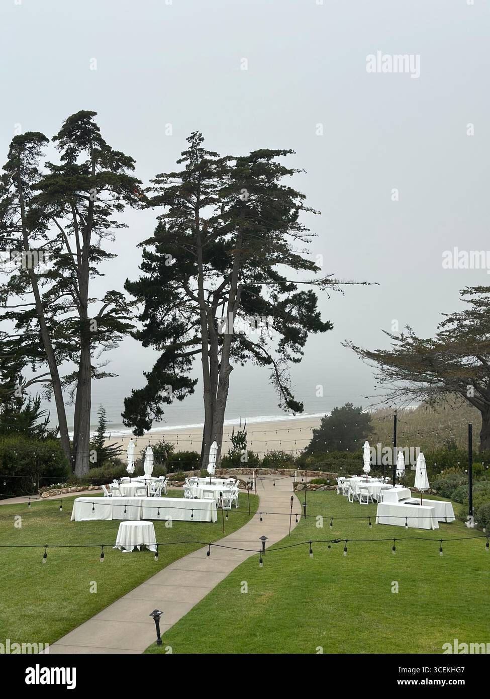 Elegant outdoor wedding and event setup with white tables, chairs, and umbrellas arranged on a lawn overlooking the Pacific Ocean on a foggy day - Smartphone Captured Stock Image