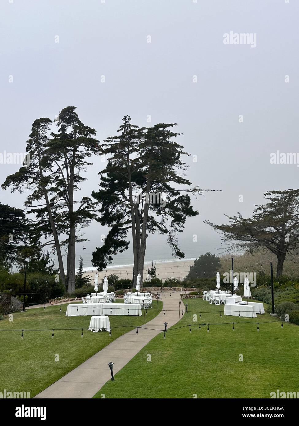 Elegant outdoor wedding and event setup with white tables, chairs, and umbrellas arranged on a lawn overlooking the Pacific Ocean on a foggy day - Smartphone Captured Stock Image