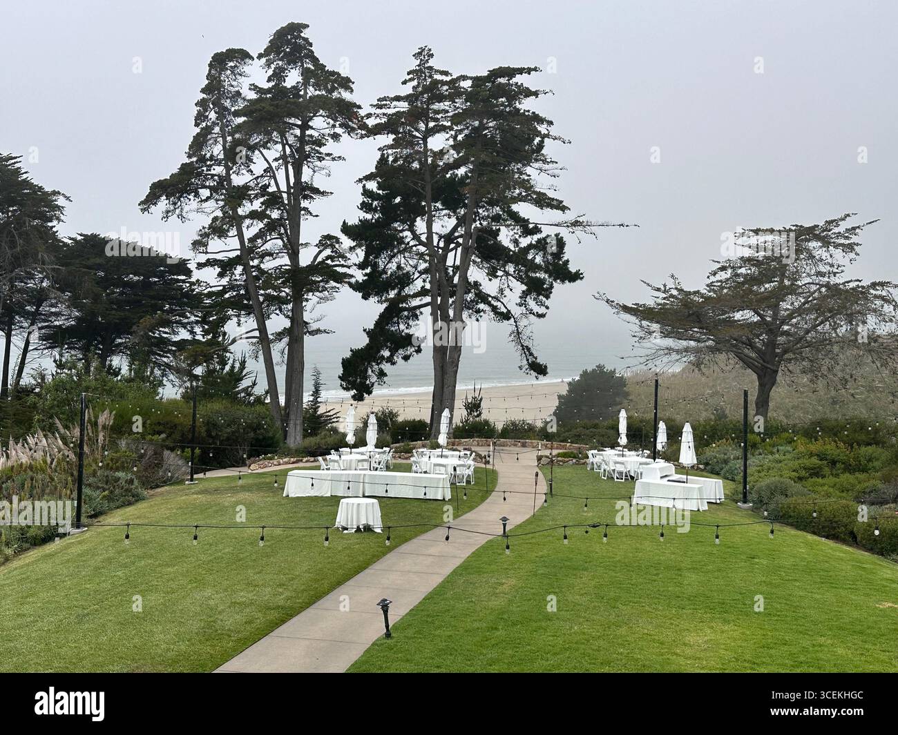 Elegant outdoor wedding and event setup with white tables, chairs, and umbrellas arranged on a lawn overlooking the Pacific Ocean on a foggy day - Smartphone Captured Stock Image