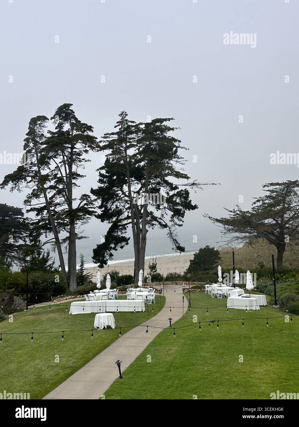 Elegant outdoor wedding and event setup with white tables, chairs, and umbrellas arranged on a lawn overlooking the Pacific Ocean on a foggy day - Smartphone Captured Stock Image