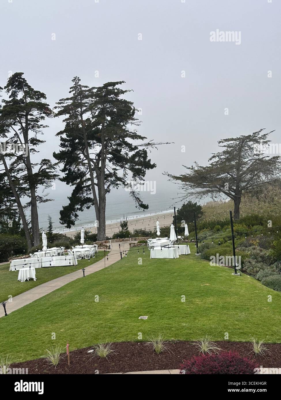 Elegant outdoor wedding and event setup with white tables, chairs, and umbrellas arranged on a lawn overlooking the Pacific Ocean on a foggy day - Smartphone Captured Stock Image