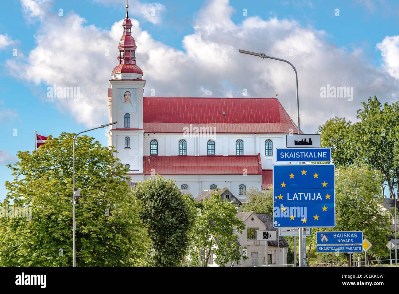 European Union border sign welcoming travelers to Latvia in Skaistkalne, with a historic church featuring a red roof and white walls in the background Stock Photo