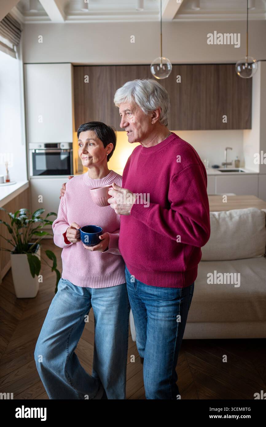 Senior couple watching street through window, talking and drinking tea. Community gossip exchange Stock Photo
