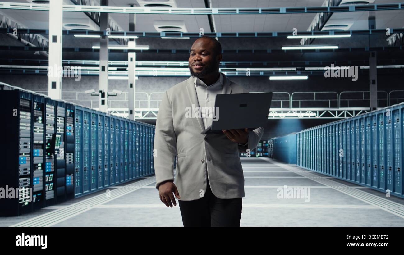 Smiling server room engineer performing maintenance on server racks using software on laptop. Happy data center IT professional upgrading blade server components using notebook, camera A Stock Photo