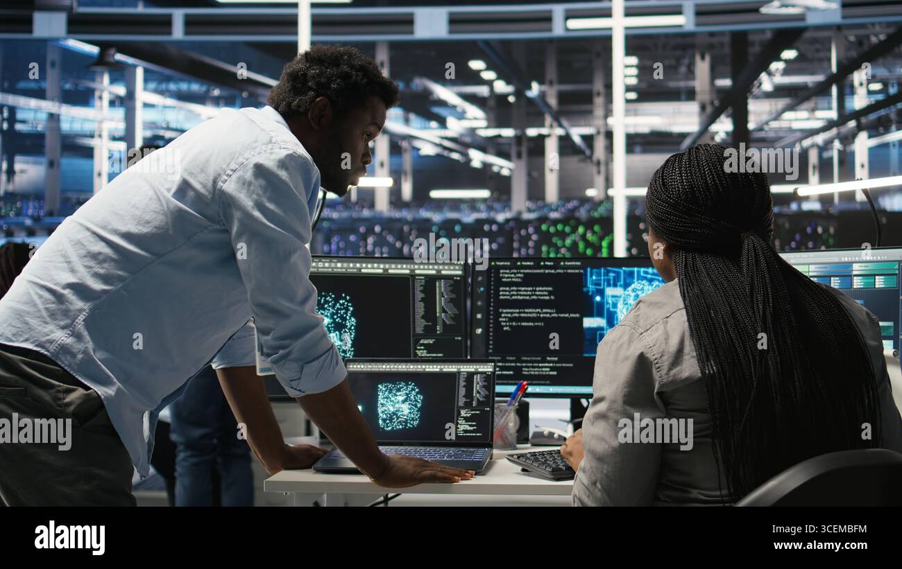 Data center engineering team uses artificial intelligence deep learning models to configure gear. Teamworking colleagues work on AI programming language in server farm, optimizing system, camera A Stock Photo