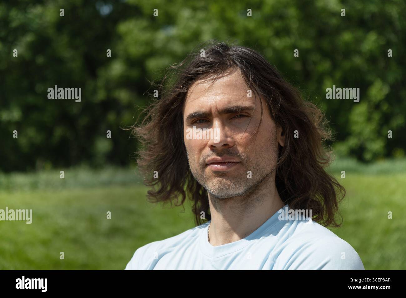 Portrait of handsome caucasian brunette man with long curly hair and dissatisfied emotion and feeling unwell at park and green nature setting. Summer Stock Photo