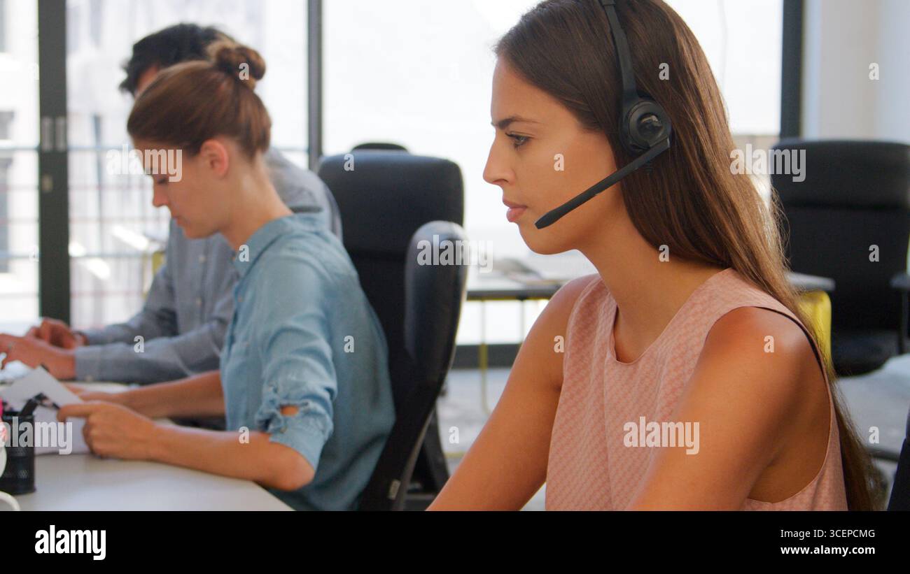 Businesswoman Wearing Headset Sitting At Desk In Office Using Computer Stock Photo