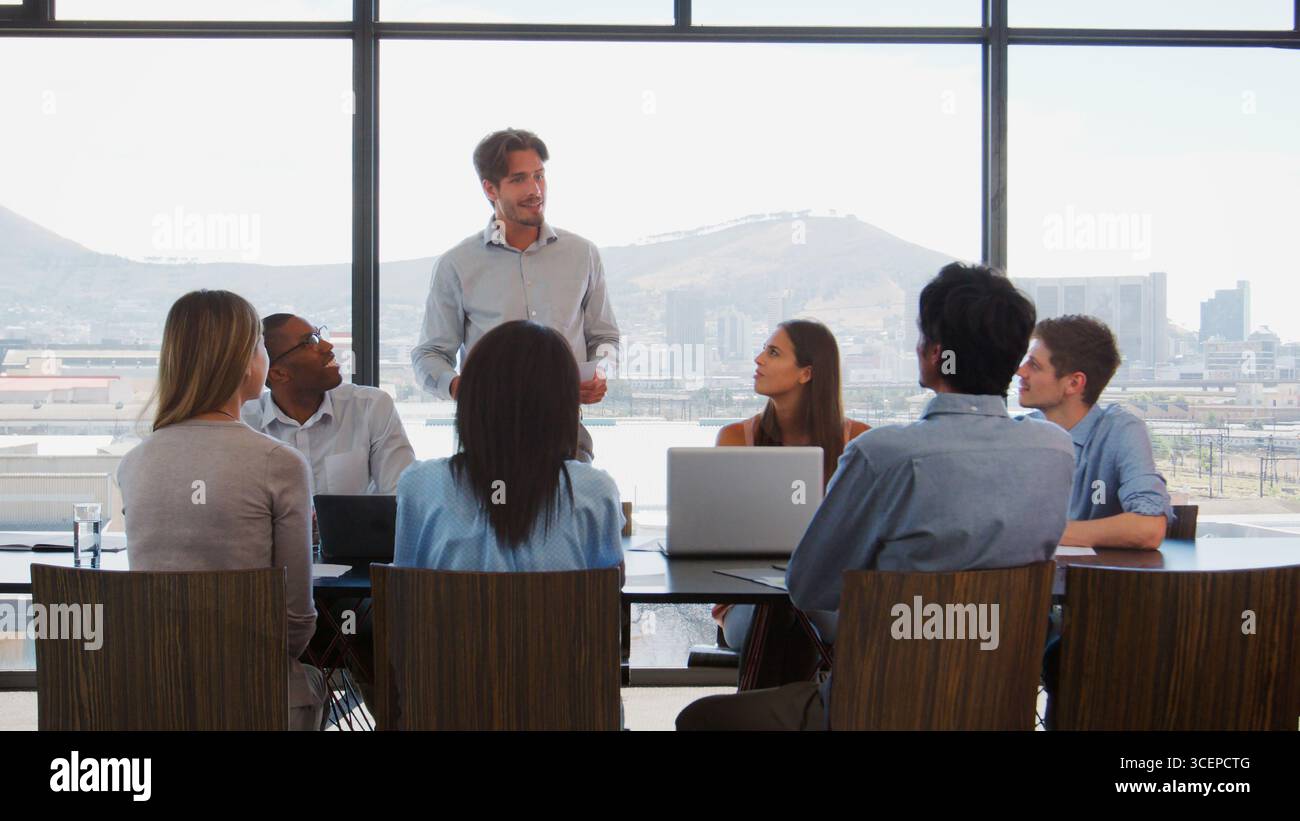 Businessman Making Presentation To Colleagues Using Laptops Sitting Around Boardroom Table Stock Photo