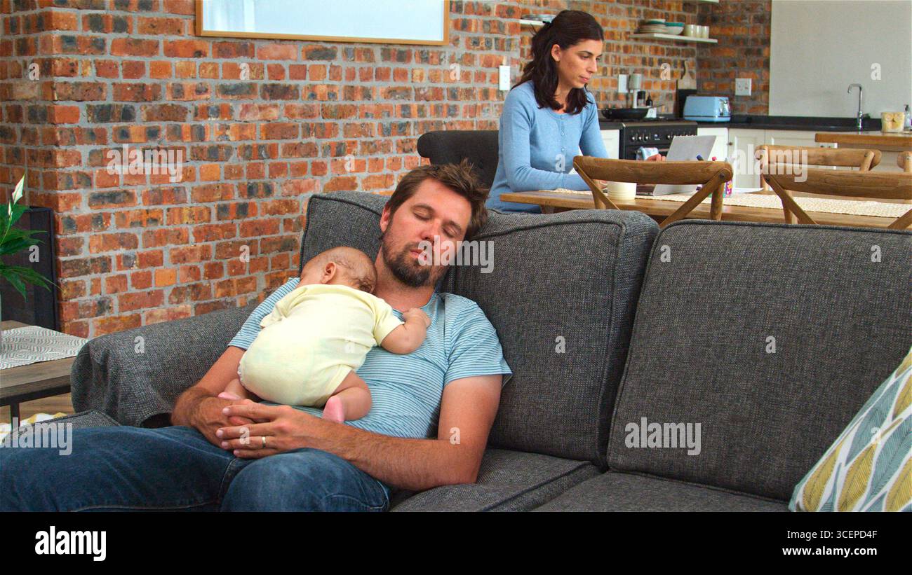 Mother Working On Laptop Whilst Tired Father Sleeps On Sofa Cuddling Baby Son Stock Photo