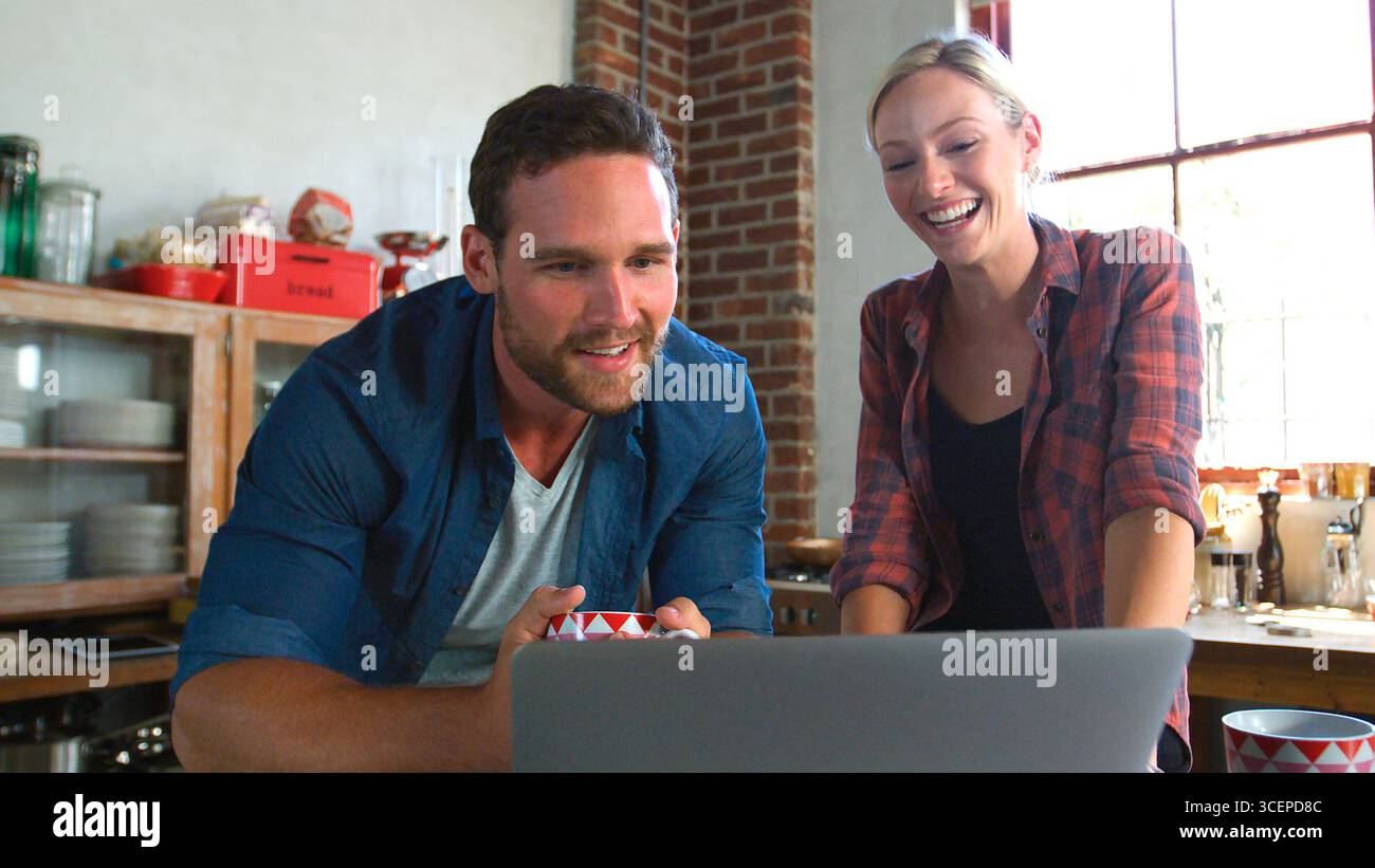 Young Couple Using Laptop In Open Plan Apartment Kitchen Stock Photo