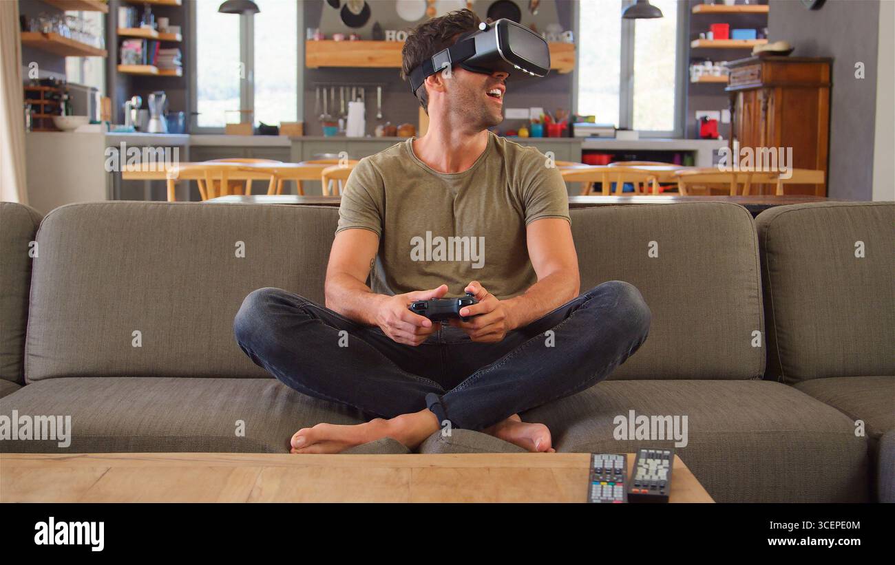 Man Sitting On Sofa At Home Playing Computer Game Using Virtual Reality Headset Stock Photo