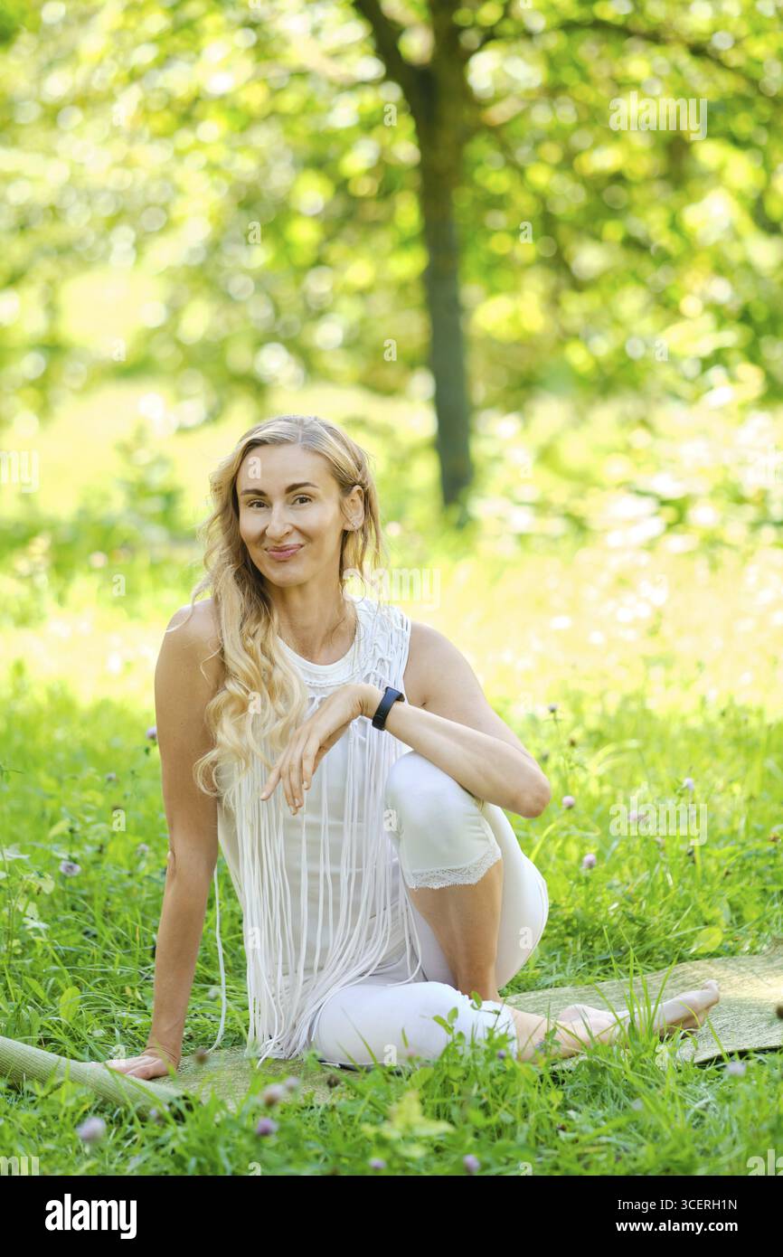 A woman dressed in white sits gracefully on a mat in a lush park ...
