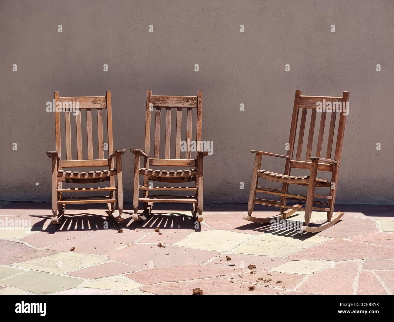 Three empty wooden rocking chairs in a row on a sunny patio; can be used conceptually, place in the sun, empty seats, trio, etc. - Smartphone Captured Stock Image