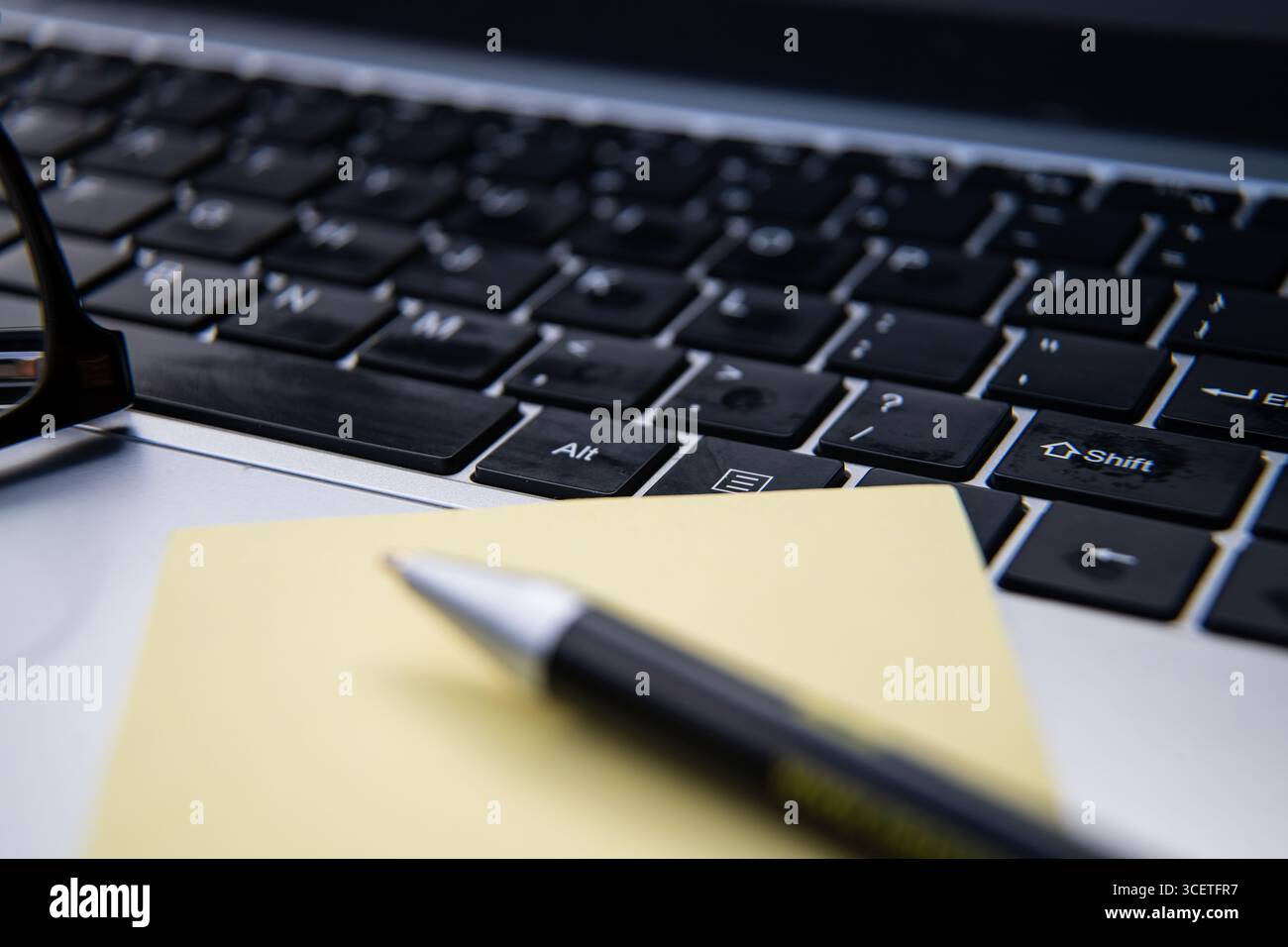 Laptop keyboard with reading glasses, pen, and yellow sticky note pad on a desk, symbolizing office work, productivity, and business planning. Stock Photo