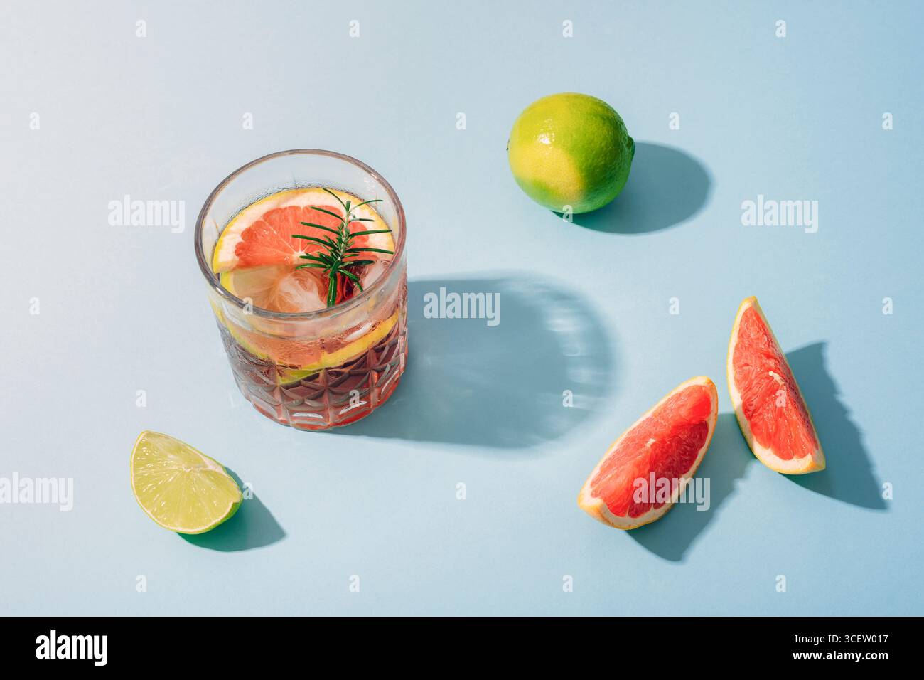 Glass of grapefruit cocktail with lime slices and rosemary on blue background with sharp shadows. Top view. Stock Photo