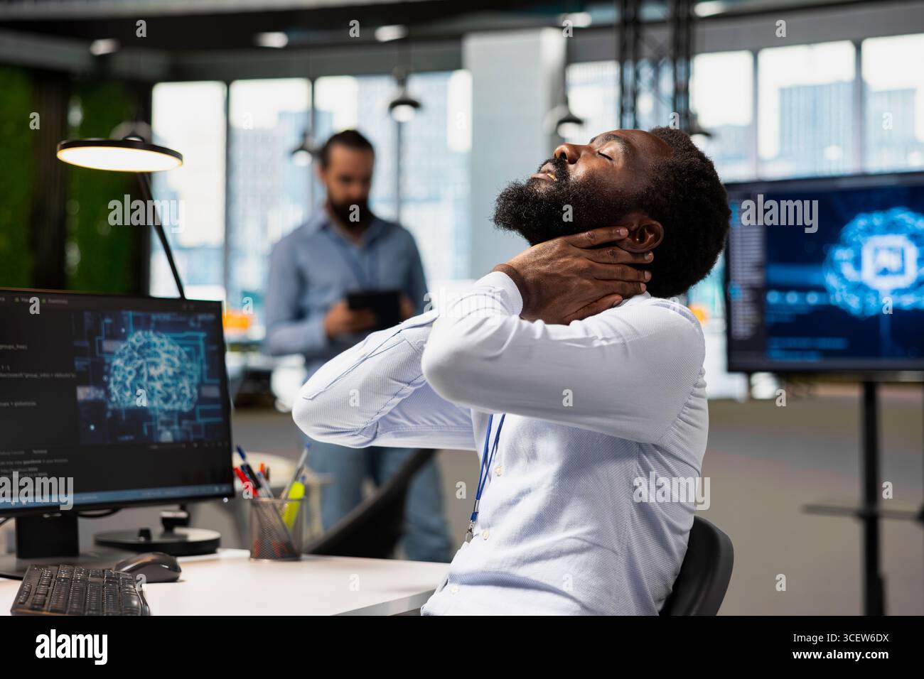 Tired engineer in office relaxing stiff neck while using AI tools to automate routine coding tasks. Fatigued IT expert massaging neck to remove pain, using artificial intelligence to do development Stock Photo