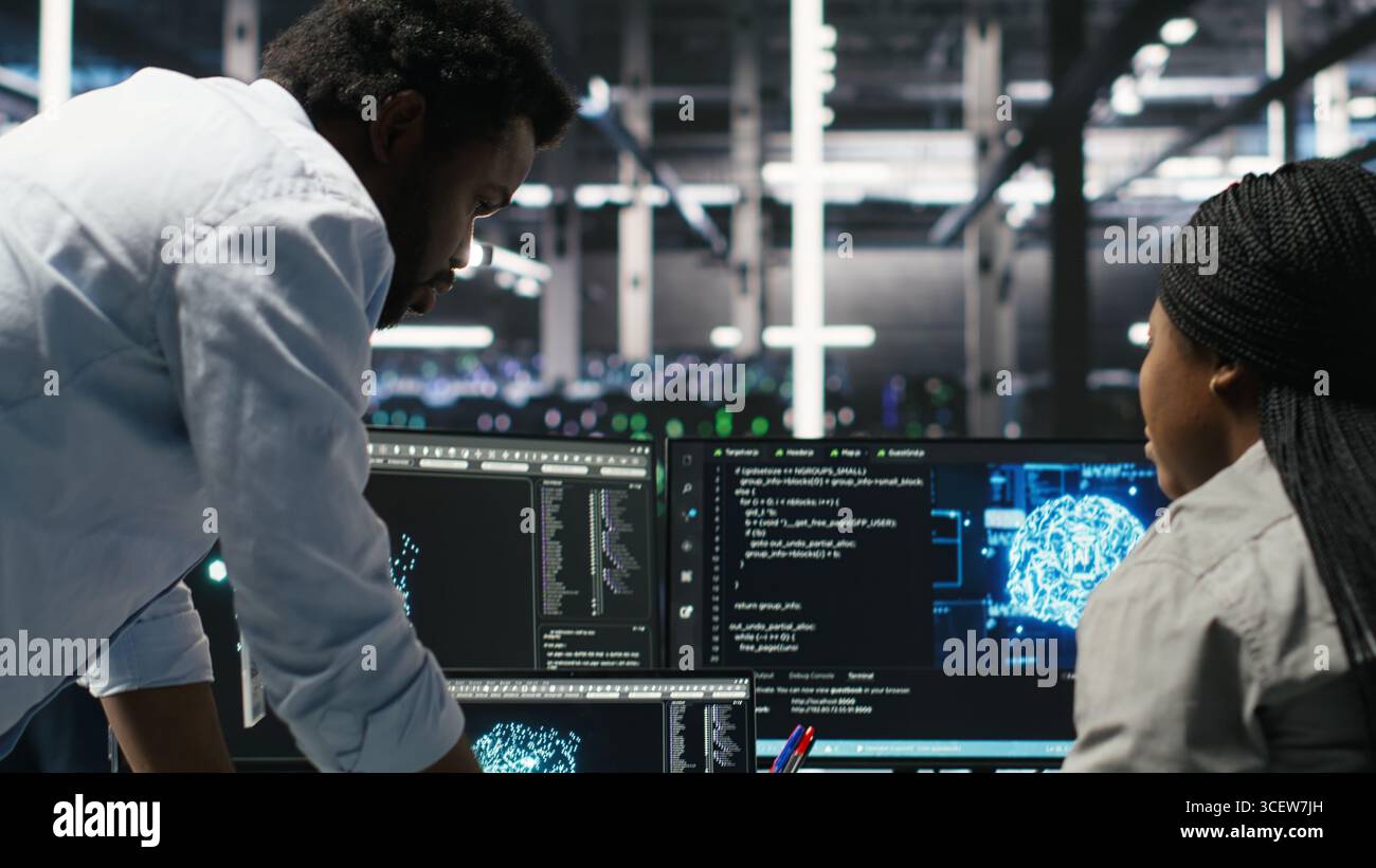 Data center engineering team uses artificial intelligence deep learning models to configure gear. Teamworking colleagues work on AI programming language in server farm, optimizing system, camera B Stock Photo