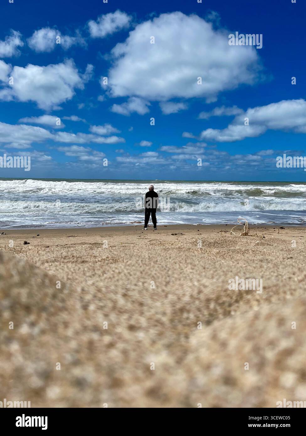 Person standing on a wide sandy beach with waves crashing under a partly cloudy sky - Smartphone Captured Stock Image
