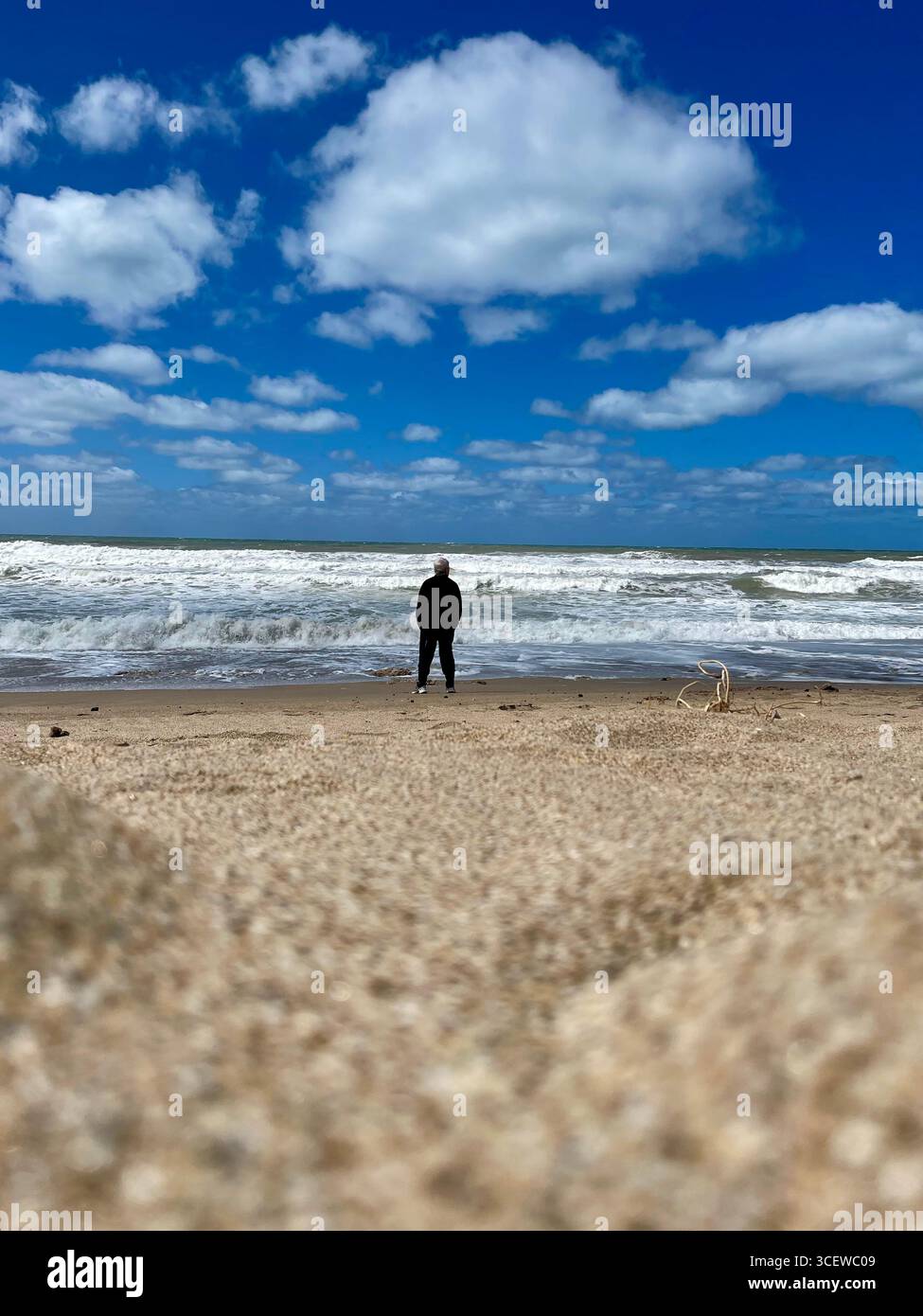 Lone person walking on a sandy beach under clear blue sky by the ocean. - Smartphone Captured Stock Image