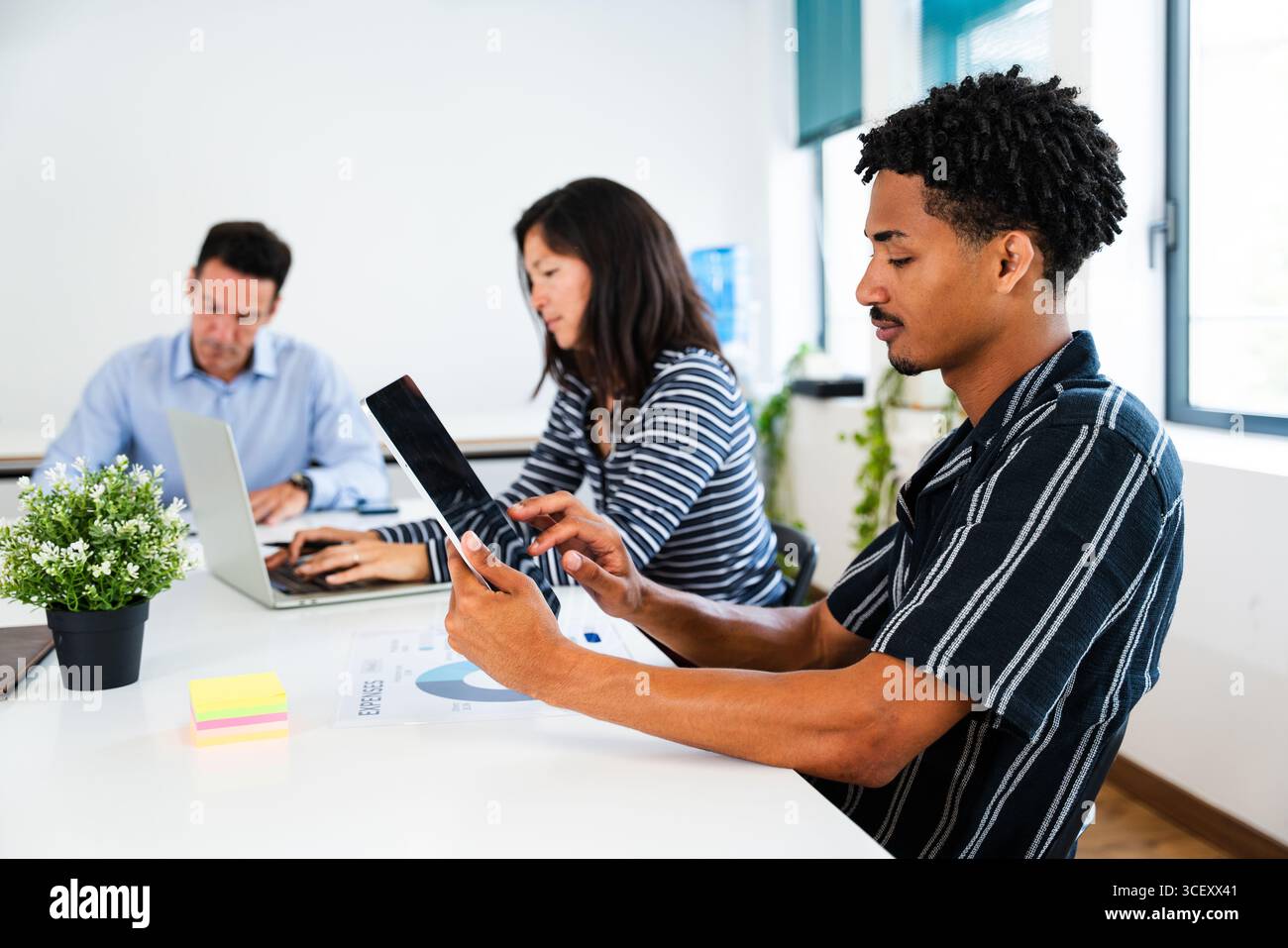 Office workers collaborating on a project at a white table, using digital devices and discussing documents, creating a dynamic and productive work env Stock Photo