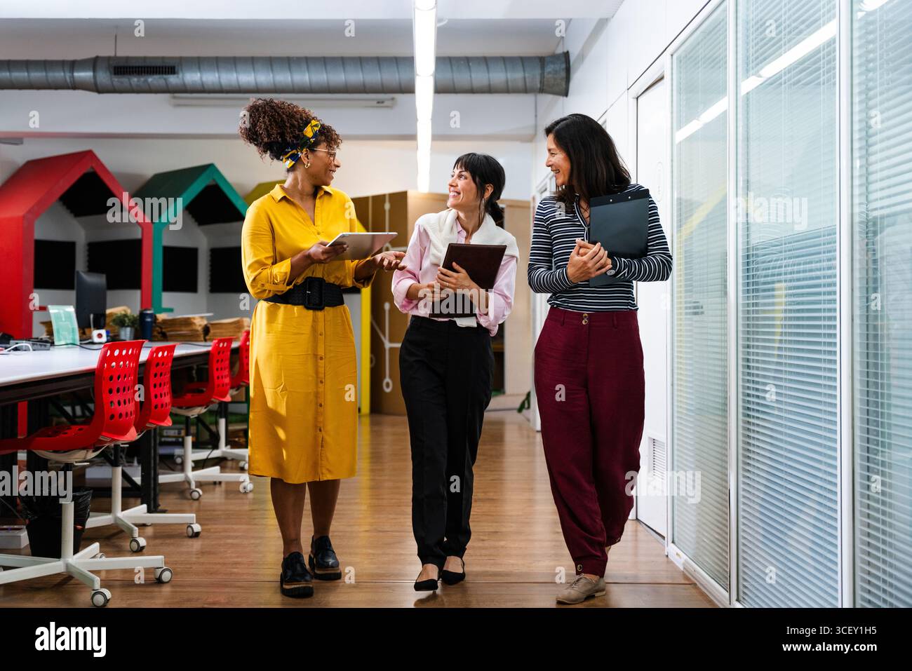 Three businesswomen are walking down an office corridor, holding digital tablets and clipboards, discussing work and smiling, showing teamwork and col Stock Photo