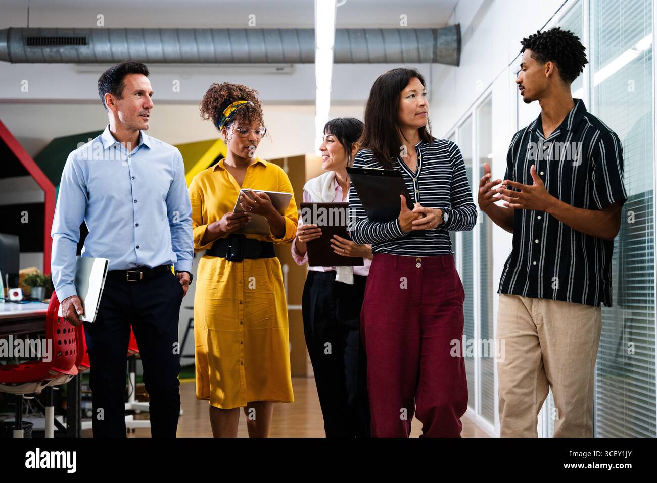 Startup business team members walking through office corridor, holding tablets, laptops, and clipboards, discussing work and collaborating on project Stock Photo