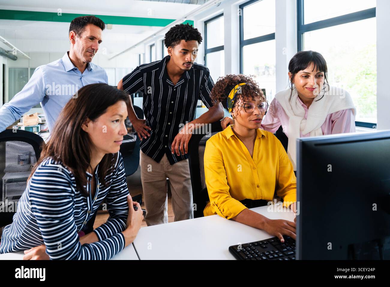 Diverse team of business professionals gathered around computer, analyzing data and discussing project in bright, modern office environment, showcasin Stock Photo