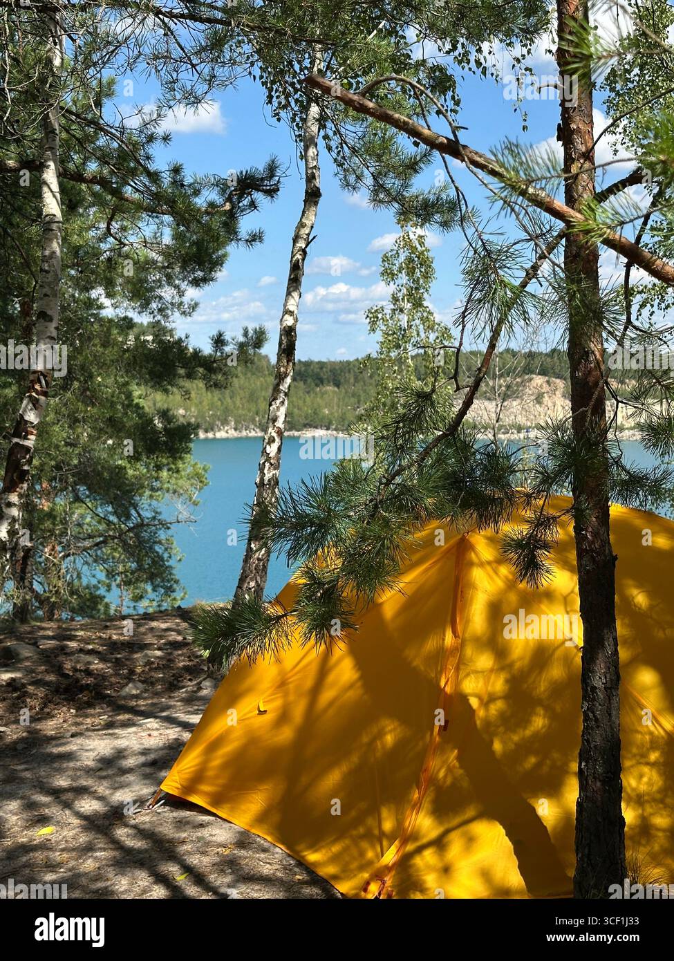 Yellow tent in forest with view of blue lake and trees. Bright yellow camping tent in a forest overlooking a turquoise blue lake with trees and cliffs - Smartphone Captured Stock Image