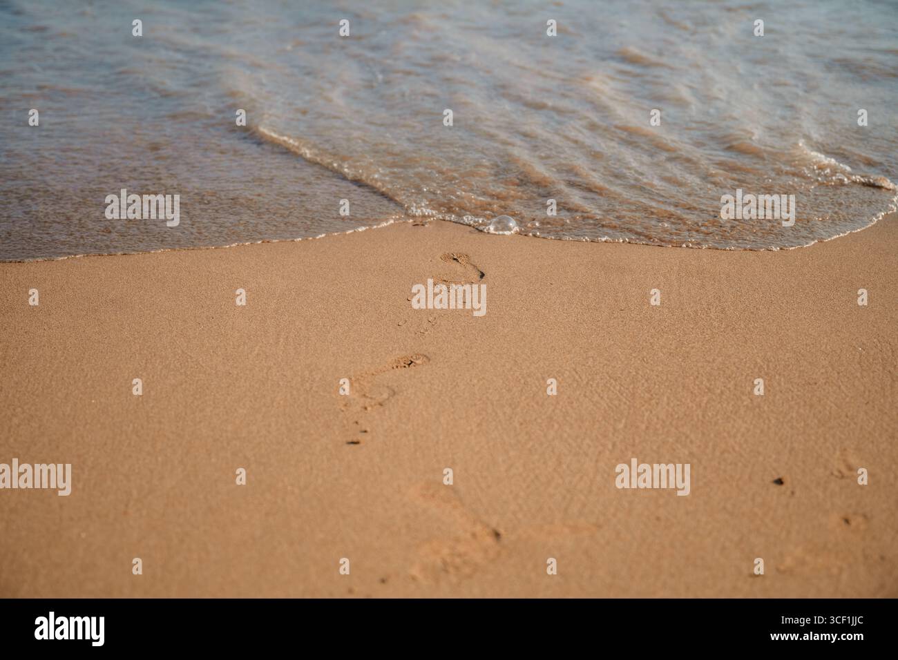 Footprints slowly disappearing beneath gentle ocean waves washing over the sandy beach, creating a tranquil scene that captures the essence of imperma Stock Photo