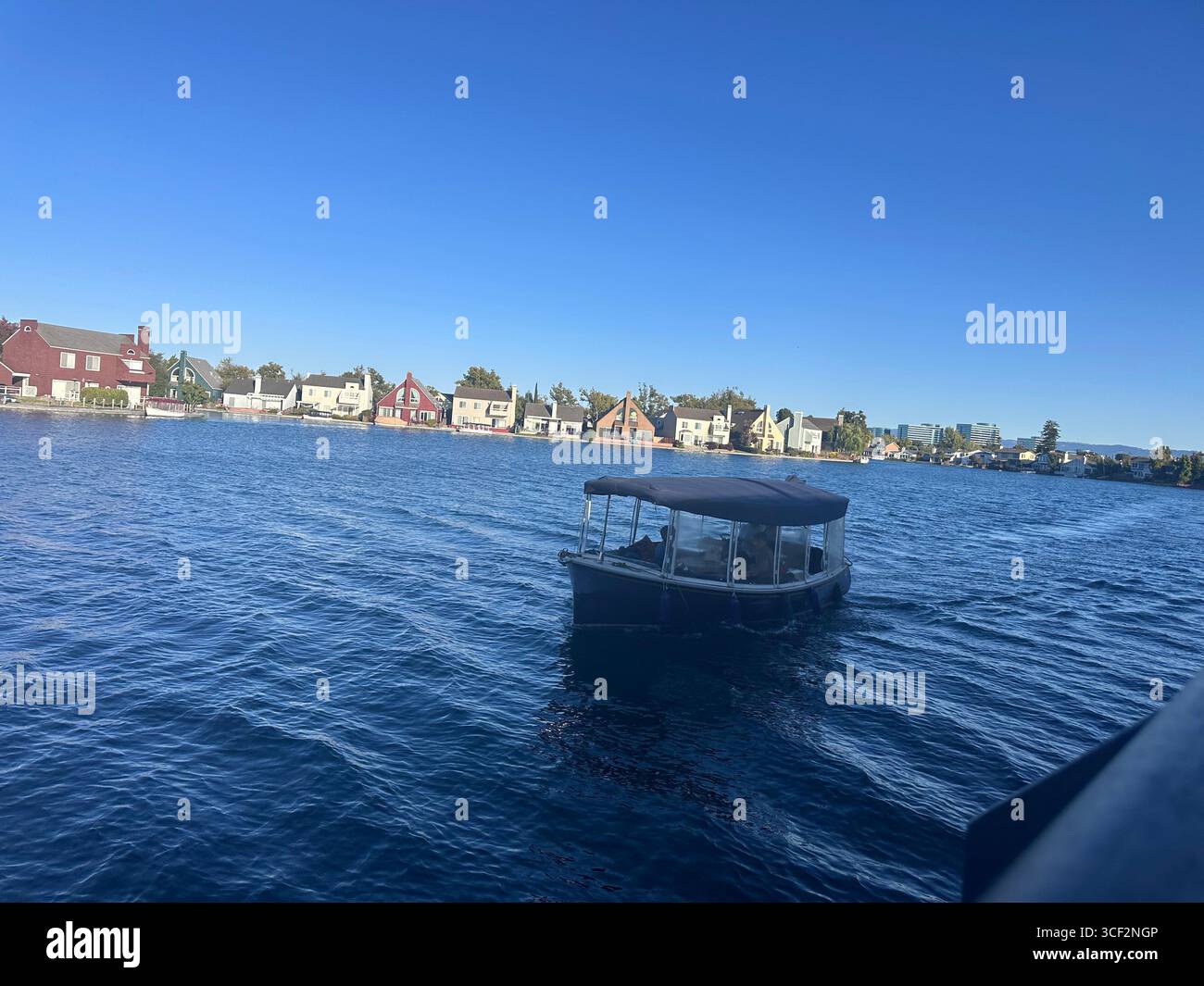 A small covered boat moves across blue water in front of colorful waterfront houses on a sunny day, with clear skies and distant modern buildings in t - Smartphone Captured Stock Image