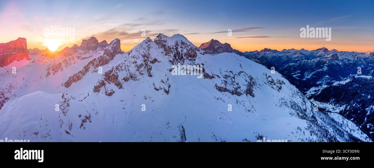 Aerial winter view of Passo Giau at dawn, with the sun rising behind Forcella Giau and Monte Cernera visible on the right. Snow-covered alpine slopes glow in the early morning light. Dolomites, Colle Santa Lucia municipality, Belluno province, Veneto region, Italy Stock Photo