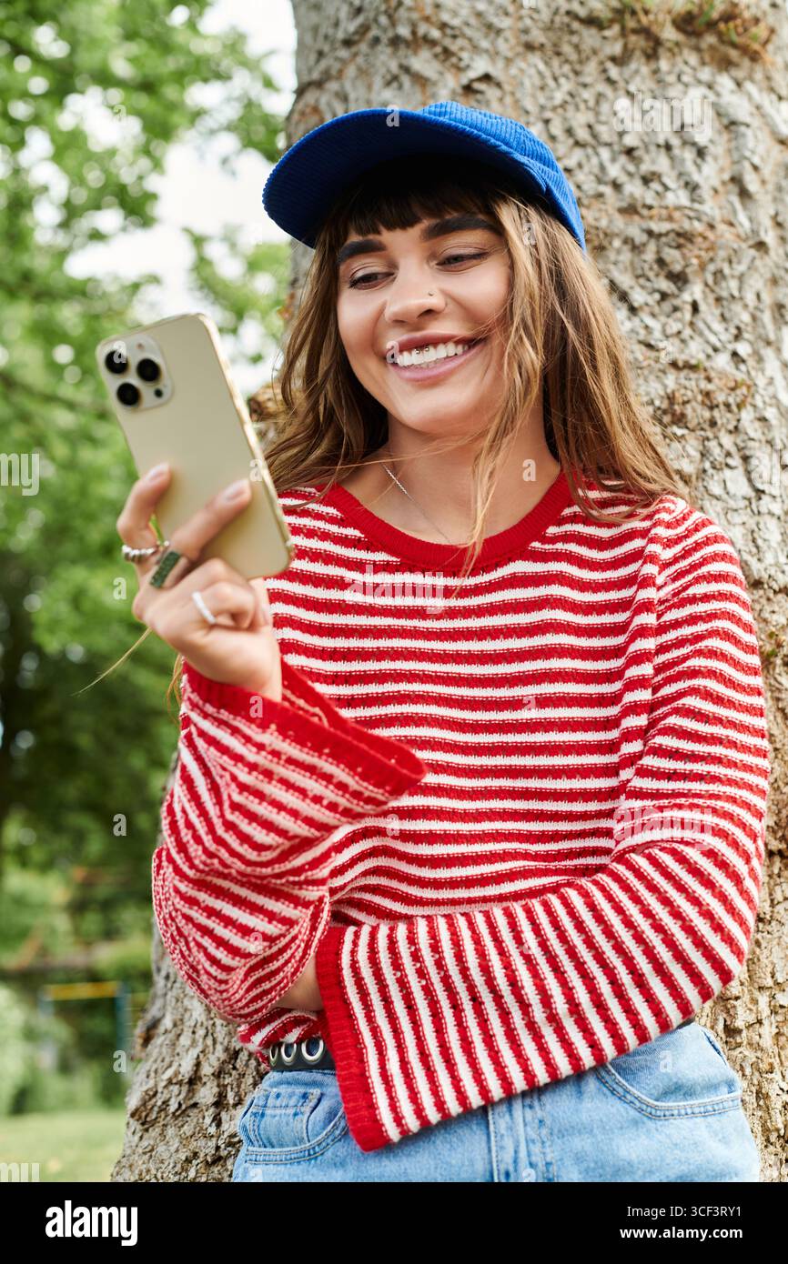 A young woman in a striped sweater smiles at her phone, leaning against a tree in the park. Stock Photo