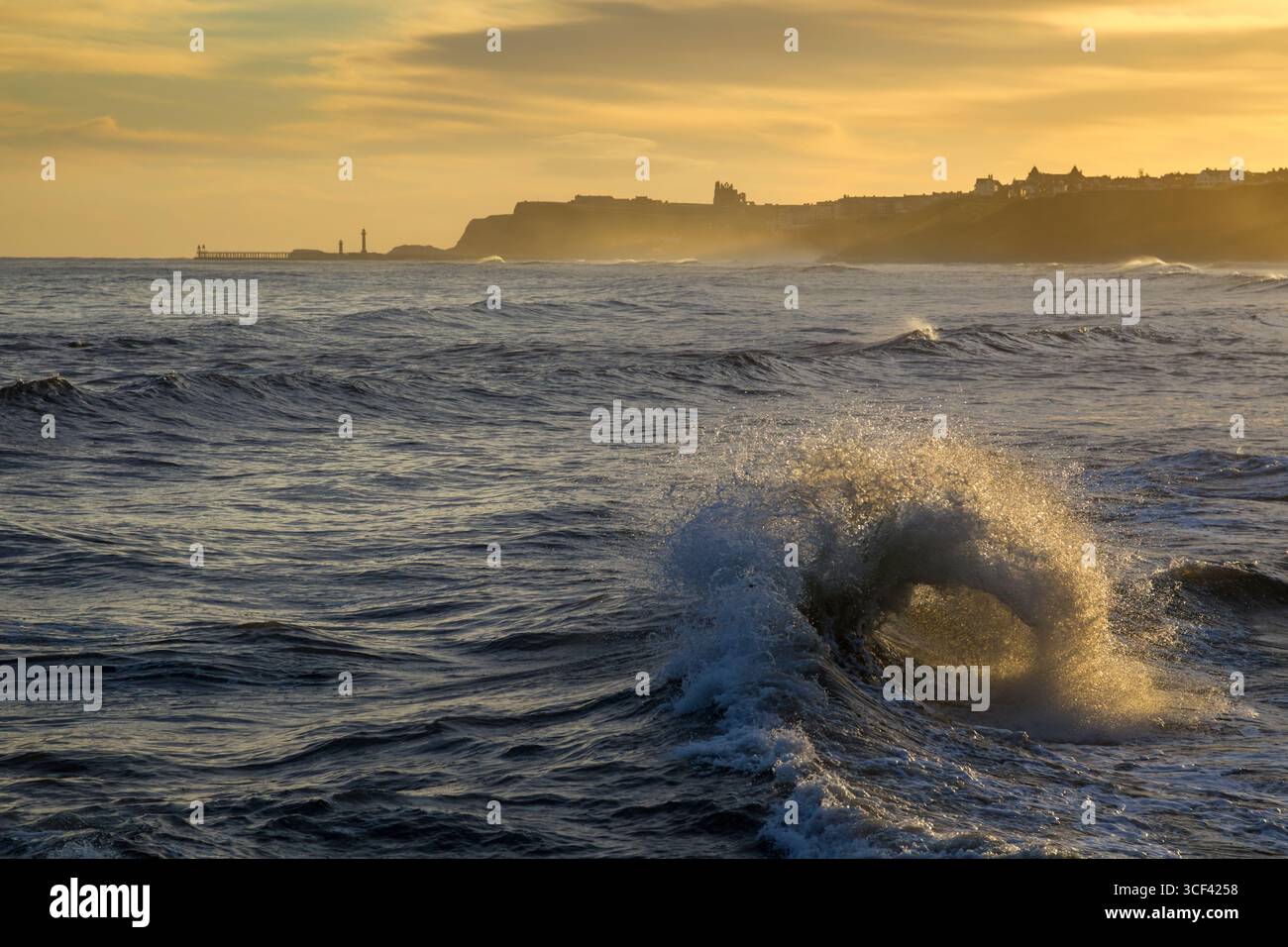 A view of a breaking wave at Sandsend looking towards the popular holiday destination and fishing port of Whitby on the northeast coast of England - Stock Image