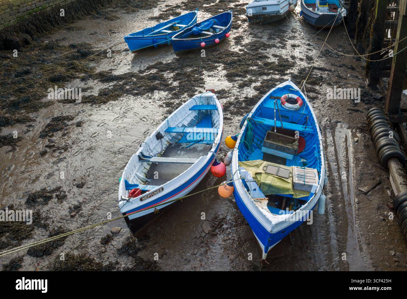 Small blue and white boats in mud moored in Whitby harbour at low tide showing a variety of boat equipment and also footprints in the mud - Stock Image