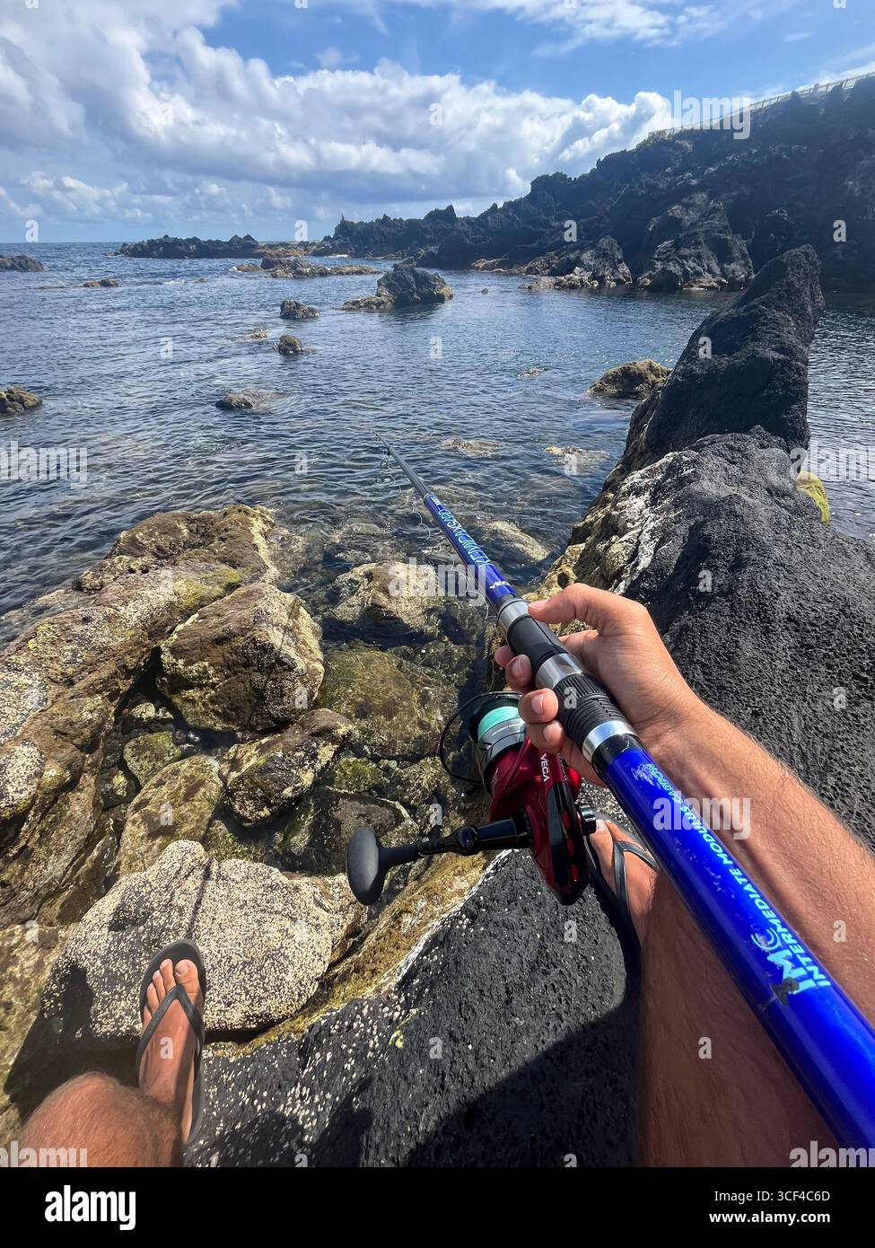 Man fishing on the rocky volcanic coastline of the Azores, Portugal, with clear Atlantic waters and dramatic seascape. - Smartphone Captured Stock Image