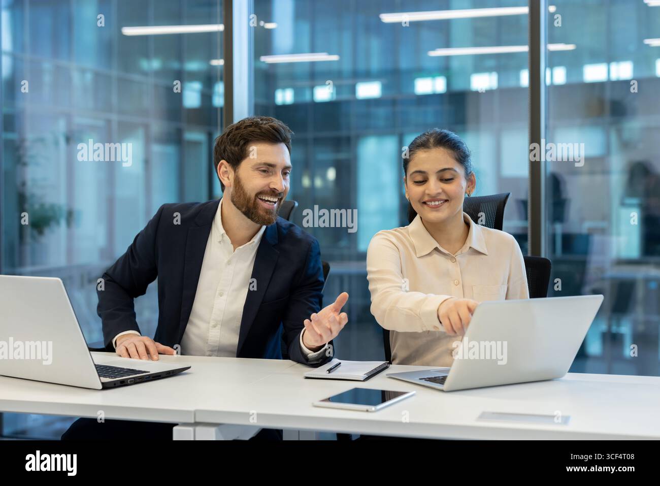 Two professional coworkers engaged in teamwork, using laptops while discussing ideas in a brightly lit office. Stock Photo