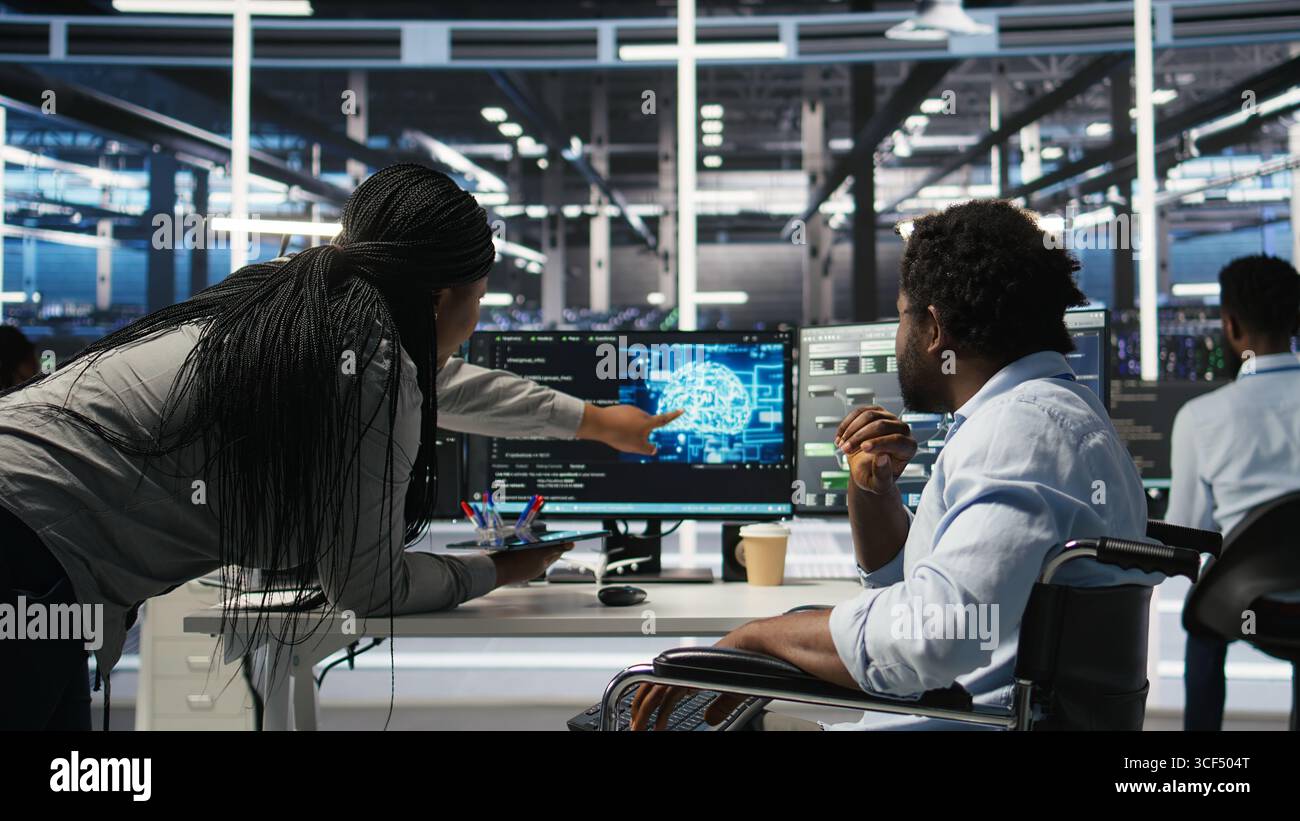 Data center engineer in wheelchair and colleague oversee automated tasks using node tree programming tool. Inclusive server farm team adjusts system settings based on AI analytics, camera A Stock Photo