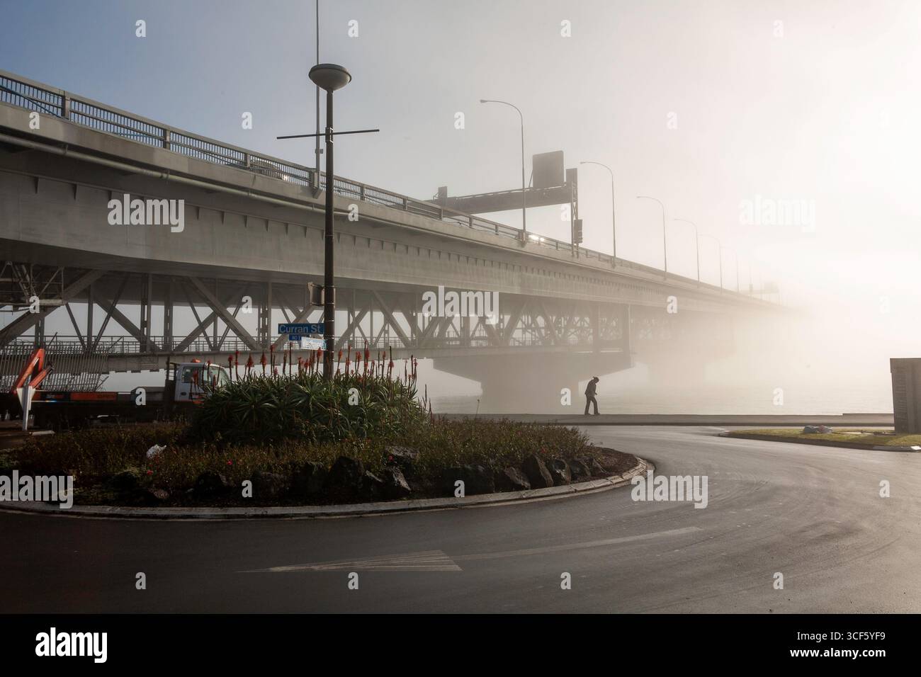 one person walking along pathway harbour side curran street westhaven drive roundabout auckland harbour bridge heavy foggy morning city fog white grey Stock Photo