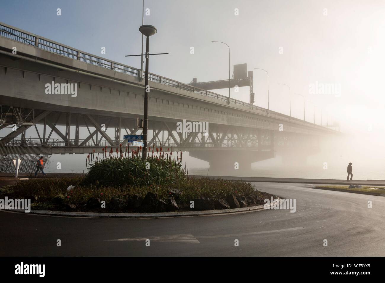 misty morning view featuring a large steel bridge over a serene river a roundabout with vegetation, two pedestrians, and urban structures creates a dy Stock Photo