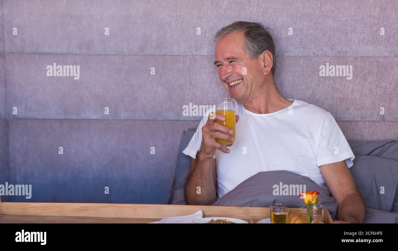 Senior man wearing white tee sitting on bed drinking orange juice near breakfast tray, copy space Stock Photo