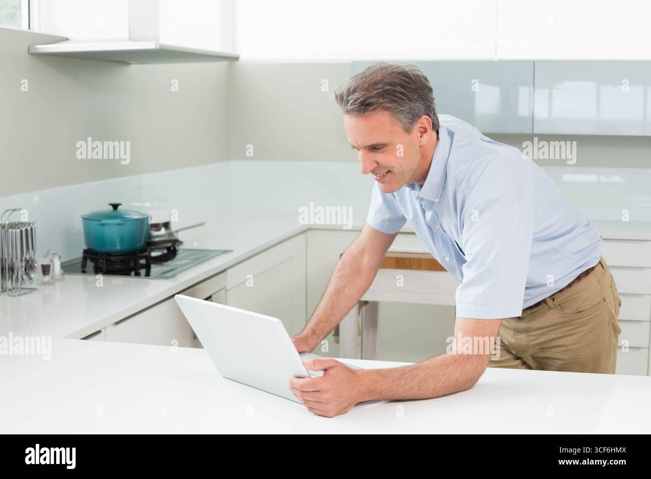 Middle-aged man leaning over countertop using white laptop beside teal pot in kitchen, copy space Stock Photo