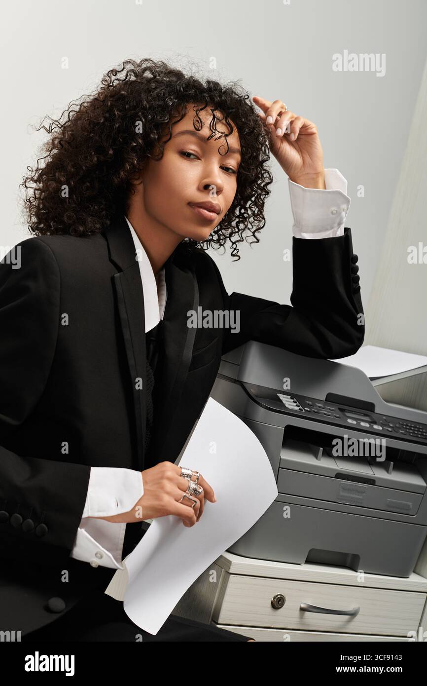 A confident young woman in a business suit poses thoughtfully by a printer. Stock Photo