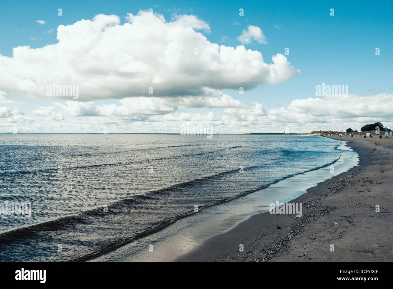 People walking along a sandy beach near the shoreline, with the water gently meeting the shore in a cloudy day Stock Photo